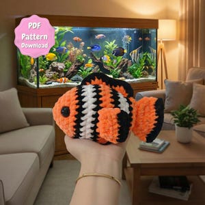 May include: A handmade, plush clownfish in orange, black, and white. The fish has a round black eye and is held in front of an aquarium filled with live fish. A pink sticker in the upper left corner says "PDF Pattern Download".