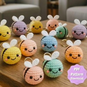 May include: A collection of colorful, crocheted bee keychains. Each bee has a smiling face, white wings, and a gold-colored keyring. The bees are in various colors, including yellow, orange, pink, blue, green, and purple. The image also includes a graphic that says "PDF Pattern Download."