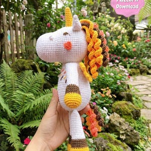 May include: A handmade white crochet unicorn with a yellow horn, orange cheeks, and a curly mane in shades of yellow, orange, and brown. The unicorn has yellow and brown hooves and a matching tail. The image includes a pink PDF pattern download graphic.