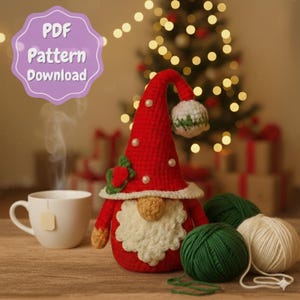 Op de afbeelding: Een handgemaakte rood-witte gehaakte kabouter met een lange, puntige hoed versierd met parels en een witte baard. De kabouter staat naast een kopje thee en bollen wol, met een kerstboom op de achtergrond. De afbeelding bevat de tekst "PDF Pattern Download."