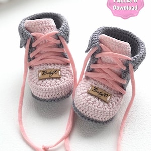 Op de afbeelding: Een paar handgehaakte baby slofjes in roze en grijs, die op kleine sneakers lijken. De slofjes hebben roze veters, een grijze kraag en een klein houten label met "Baby" en een hartje.