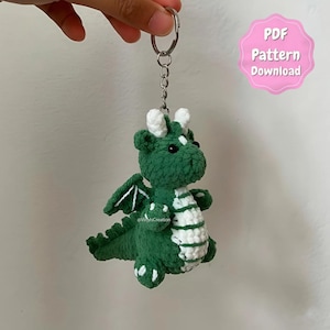May include: A green and white crocheted dragon keychain with a silver keyring. The dragon has white accents on its horns, belly, wings, and feet. The keychain is a handmade item. The image also includes a pink tag that says "PDF Pattern Download."