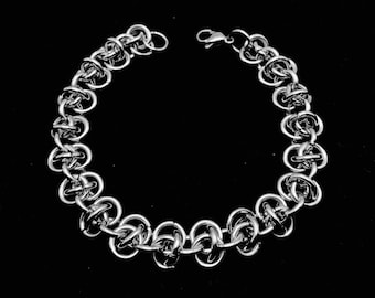 May include: A silver chain bracelet with a unique interlocking design. The bracelet is made of small, round links that are woven together to create a intricate pattern.