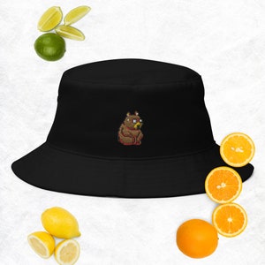 Puede incluir: Sombrero de pescador negro con un oso pixelado marrón con gafas bordadas en la parte delantera.