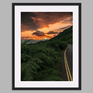 May include: A winding road with yellow lines leads through a lush green forest towards a mountain range with a bright orange sunset in the background.