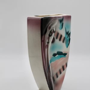 May include: A rectangular ceramic vase with a cream-colored front and a design on the side. The design features a face-like shape with pink, teal, and black accents. The vase has a square opening at the top.