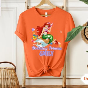 Peut inclure: T-shirt orange avec un motif color&eacute; de sir&egrave;ne aux cheveux roux et &agrave; la queue verte. Le texte "Birthday Princess & EMILY" est imprim&eacute; en dessous. Le t-shirt est suspendu &agrave; un portant en bois.