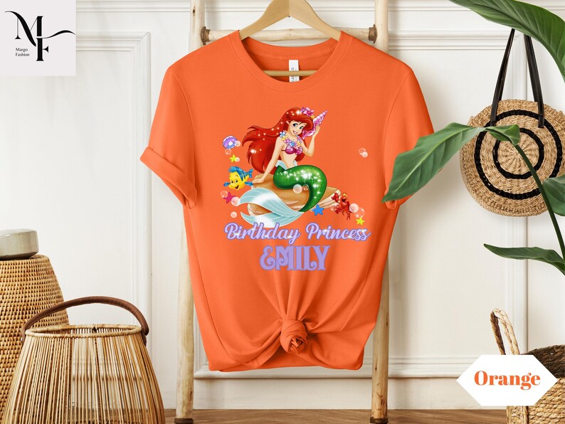 Peut inclure: T-shirt orange avec un motif color&eacute; de sir&egrave;ne aux cheveux roux et &agrave; la queue verte. Le texte "Birthday Princess & EMILY" est imprim&eacute; en dessous. Le t-shirt est suspendu &agrave; un portant en bois.