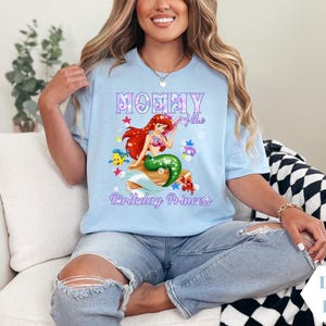Peut inclure: T-shirt bleu clair avec un motif de sir&egrave;ne, de poissons et le texte "Mommy of the Birthday Princess". Le t-shirt est de couleur bleu clair.