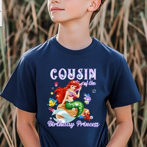 Peut inclure: T-shirt bleu marine avec un motif de sir&egrave;ne aux cheveux rouges et &agrave; la queue verte, entour&eacute;e de cr&eacute;atures marines. Le texte "Cousin of the Birthday Princess" est affich&eacute; au-dessus et en dessous du motif.
