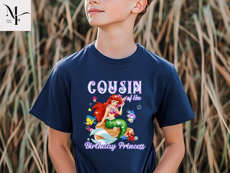 Peut inclure: T-shirt bleu marine avec un motif de sir&egrave;ne aux cheveux rouges et &agrave; la queue verte, entour&eacute;e de cr&eacute;atures marines. Le texte "Cousin of the Birthday Princess" est affich&eacute; au-dessus et en dessous du motif.