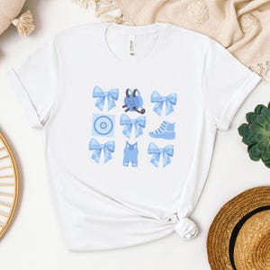 May include: White t-shirt with a blue graphic design featuring a variety of blue bows, a shoe, a cat, a target, and a creature with horns.