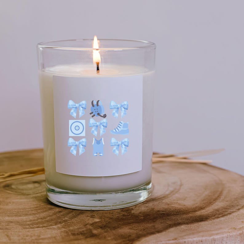 May include: A clear glass candle jar with a white candle and a blue and white label. The label has a variety of baby-themed icons, including a pacifier, a rattle, a pair of shoes, and a onesie.
