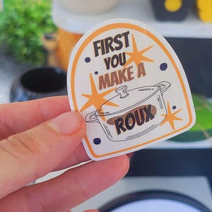 May include: White sticker with a yellow border and black text that reads "First you make a roux". The sticker features a cartoon illustration of a pot with a lid and the word "roux" written inside the pot.
