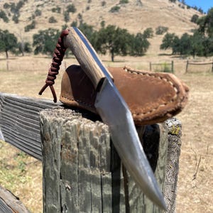 K&ouml;nnte beinhalten: Ein handgemachtes Messer mit einem Holzgriff und einer Lederscheide. Das Messer liegt auf einem Holzpfosten eines Zauns mit einem grasbewachsenen Feld und H&uuml;geln im Hintergrund.