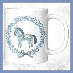 May include: White ceramic mug featuring a blue horse design within a wreath of blue leaves and berries. The horse has a patterned saddle and is surrounded by a blue bow. The mug has a curved handle.