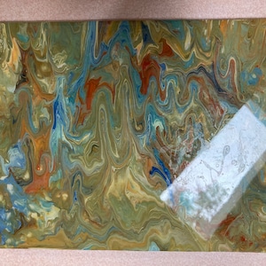 May include: Abstract acrylic painting with a fluid, marbled effect. The artwork features a blend of colors including olive green, blue, orange, and cream, creating a dynamic and textured visual. The painting is on a rectangular canvas.