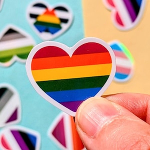 Könnte beinhalten: Eine Sammlung herzförmiger Aufkleber mit verschiedenen LGBTQ+-Pride-Flaggen-Designs. Ein Aufkleber, der von einer Hand gehalten wird, zeigt die Regenbogenflagge. Andere Aufkleber zeigen Flaggen, die verschiedene Identitäten repräsentieren, mit leuchtenden Farben und glänzendem Finish.