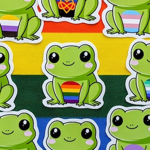 May include: A selection of green frog stickers, each displaying a different LGBTQ+ pride flag design, set against a rainbow backdrop. The stickers depict smiling frogs with large black eyes and white bellies.