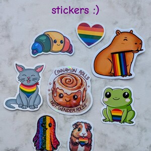 May include: A selection of colourful stickers featuring cartoon animals and food. Stickers include a croissant, heart, capybara, cat, cinnamon roll, ghost, guinea pig, and frog, all with rainbow accents. Text on the stickers reads "Cinnamon Rolls" and "Not Gender Roles."