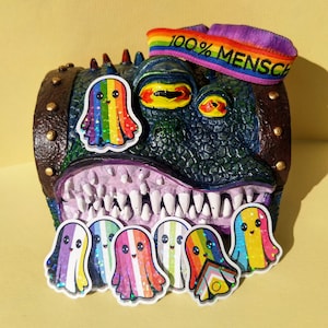 May include: A quirky, multi-coloured monster sculpture with a wide mouth and many teeth. The monster has a rainbow-coloured ghost sticker on its head and several ghost stickers in front. A rainbow headband with the text "100% MENSCH" is on top.