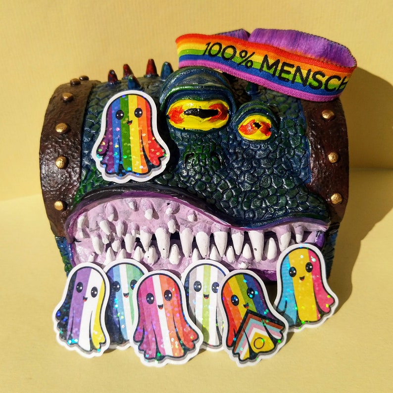 May include: A quirky, multi-coloured monster sculpture with a wide mouth and many teeth. The monster has a rainbow-coloured ghost sticker on its head and several ghost stickers in front. A rainbow headband with the text "100% MENSCH" is on top.