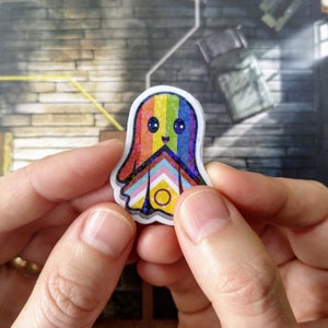 May include: A colourful sticker of a friendly ghost with a rainbow design. The ghost has a happy face and a glittery finish. The design incorporates the colours of the LGBTQ+ pride flag.