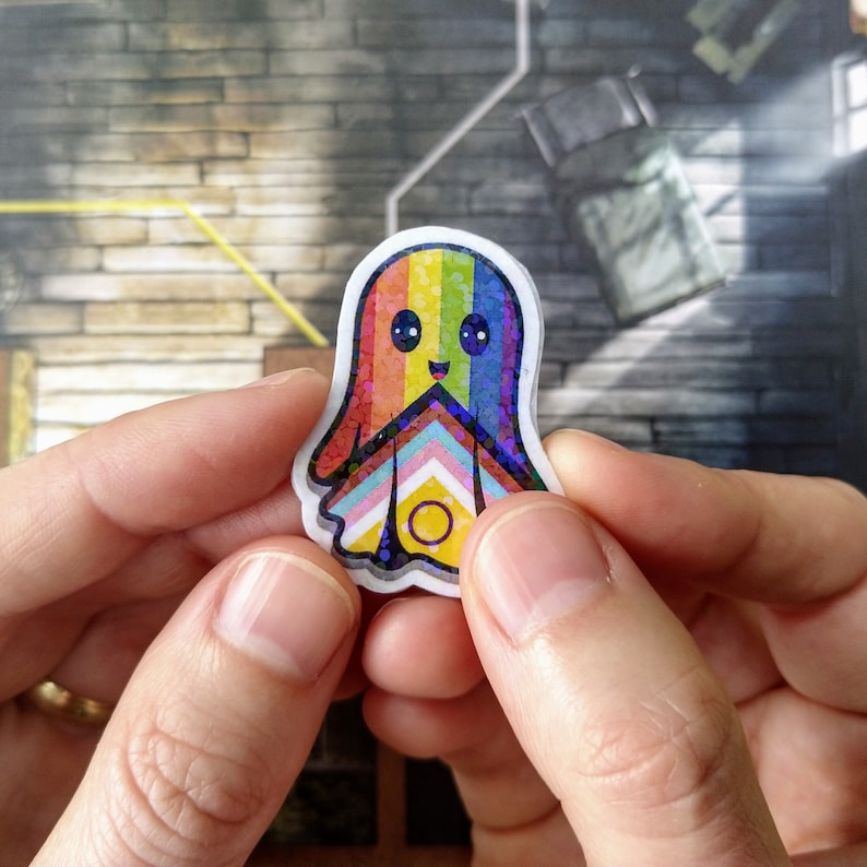 May include: A colourful sticker of a friendly ghost with a rainbow design. The ghost has a happy face and a glittery finish. The design incorporates the colours of the LGBTQ+ pride flag.
