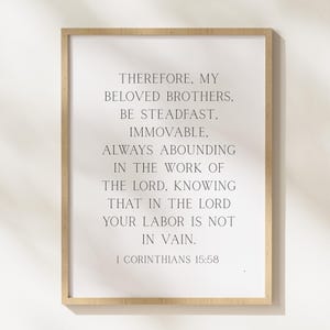 May include: A framed print with a light wood frame. The print features the text "THEREFORE, MY BELOVED BROTHERS, BE STEADFAST, IMMOVABLE, ALWAYS ABOUNDING IN THE WORK OF THE LORD, KNOWING THAT IN THE LORD YOUR LABOR IS NOT IN VAIN. 1 CORINTHIANS 15:58."
