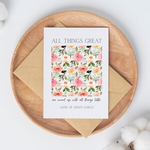 May include: A greeting card with a floral pattern of pink and peach roses, green leaves, and yellow flowers. The card is on a wooden tray with a brown envelope. Text on the card reads "ALL THINGS GREAT are wound up with all things little. ANNE OF GREEN GABLES."