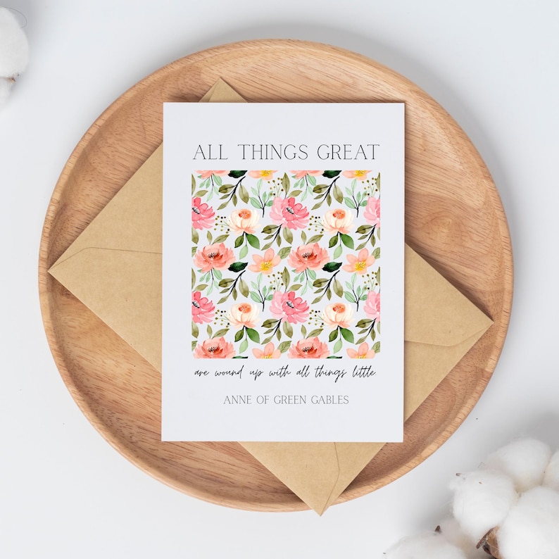 May include: A greeting card with a floral pattern of pink and peach roses, green leaves, and yellow flowers. The card is on a wooden tray with a brown envelope. Text on the card reads "ALL THINGS GREAT are wound up with all things little. ANNE OF GREEN GABLES."