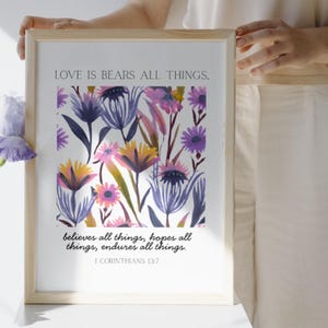 May include: A framed art print featuring a floral watercolor design in shades of purple, pink, and yellow. The text "LOVE IS BEARS ALL THINGS" is at the top, with a Bible verse below. The frame is light-colored wood.