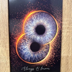 May include: A framed print showing two overlapping blue irises with black pupils, surrounded by a golden ring and fiery orange particles. The words "Always & forever" are written in cursive. The frame is made of light-coloured wood.