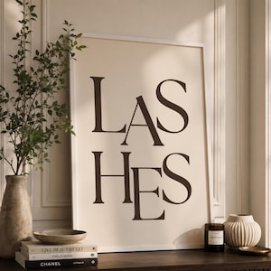 May include: A rectangular wall art print in a white frame featuring the word LASHES in large, dark brown serif typography against a light beige background. The print is displayed on a surface alongside a tall vase, books, and a candle.