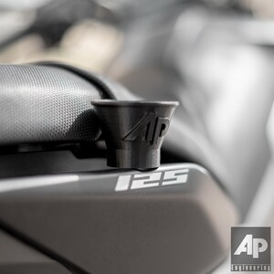May include: A black plastic motorcycle phone mount with the letters "AP" printed on it. The mount is attached to a motorcycle seat with the number "125" printed on it.