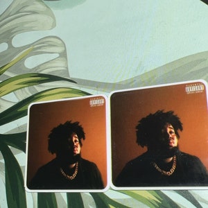 May include: Two album covers featuring a man with dark curly hair and a beard wearing a black shirt and a gold chain. The background is a solid orange color. The album covers have a parental advisory sticker with the text "Parental Advisory Explicit Content".