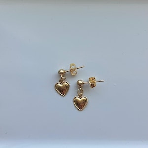 May include: A pair of gold-colored heart-shaped dangle earrings. Each earring features a small gold ball stud and a heart pendant with a textured border. The earrings are displayed on a white surface.