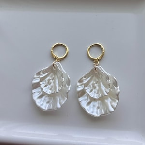 May include: A pair of white, layered petal-shaped earrings with gold-colored hoops. The earrings have a pearlescent finish and are displayed on a white surface.