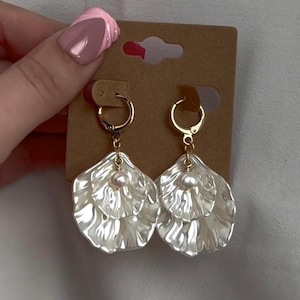 May include: A pair of white, layered petal-shaped earrings with gold-colored hoops and a small pearl at the center of each earring. The earrings are displayed on a brown earring card.