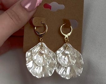 May include: A pair of white, layered petal-shaped earrings with gold-colored hoops and a small pearl at the center of each earring. The earrings are displayed on a brown earring card.