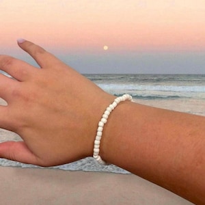 May include: A white beaded bracelet is worn on a wrist, set against a beach scene with a sunset sky. The bracelet is made of small, round, white beads strung together. The background features the ocean, sand, and a colorful sky.