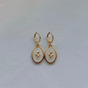 May include: A pair of gold hoop earrings featuring oval-shaped pendants. Each pendant has a cream-colored enamel inlay with a raised gold flower design in the center and a scalloped gold border surrounding the oval frame.
