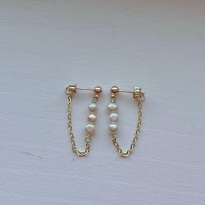 May include: A pair of gold-colored chain earrings. Each earring features a small gold ball stud, three white pearls, and a delicate gold chain. The earrings are displayed on a white surface.