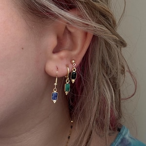 May include: Three gold-toned earrings worn in a single ear. The first is a drop earring with a rectangular blue stone, the second is a small hoop with a rectangular green stone, and the third is a stud earring featuring a rectangular black stone.