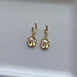 May include: A pair of gold-colored hoop earrings. Each earring features a small, white pearl suspended from a gold-colored, shell-shaped charm. The earrings are displayed on a white surface.