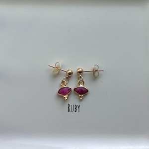 May include: A pair of gold stud earrings featuring a small round ball post and a dangling marquise-shaped faceted ruby gemstone with three tiny gold beads at the base. The word RUBY is printed in black text below the earrings.