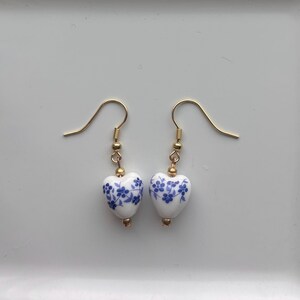 May include: A pair of drop earrings featuring white heart-shaped beads with a blue floral pattern. The beads are suspended from gold-toned metal fishhook wires, accented with small gold-toned spacer beads above and below each heart-shaped charm.