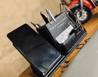 May include: Black leather desk organizer with a phone stand, pen holder, and card slots. The organizer has a personalized inscription that reads "Grateful for your contributions! Love Emma".