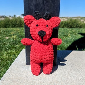 May include: A red crocheted stuffed animal with black horns and a black nose. The animal is standing on a gray concrete surface.