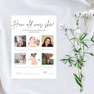 May include: A white bridal shower game card with the text "How old was she?" and six photo slots. The card includes fill-in-the-blank lines for the name and total score. White flowers and greenery are in the background.